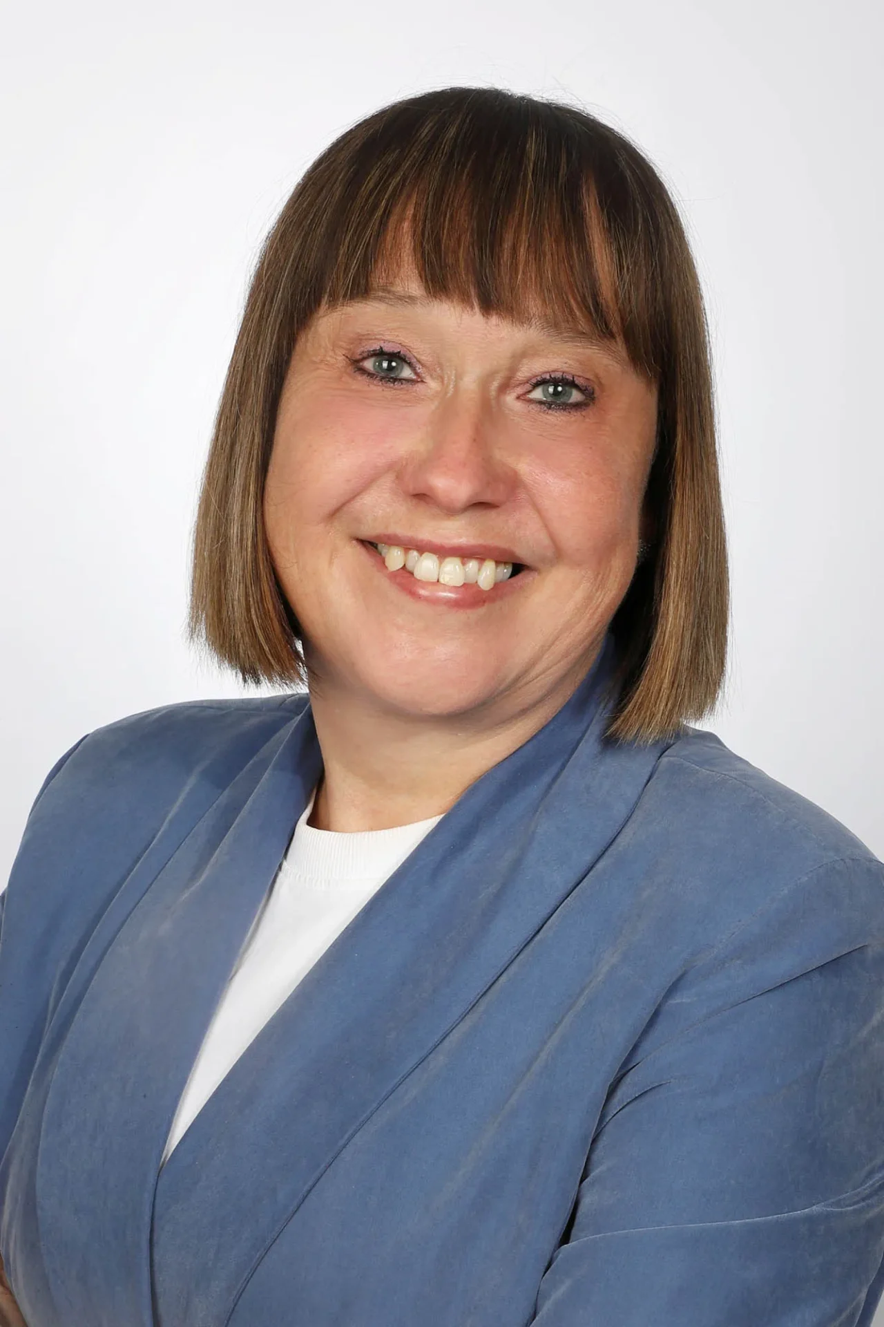 Eine Frau mit glattem braunem Haar und Pony, die einen blauen Blazer und ein weißes Oberteil trägt, lächelt in die Kamera vor einem schlichten hellen Hintergrund.