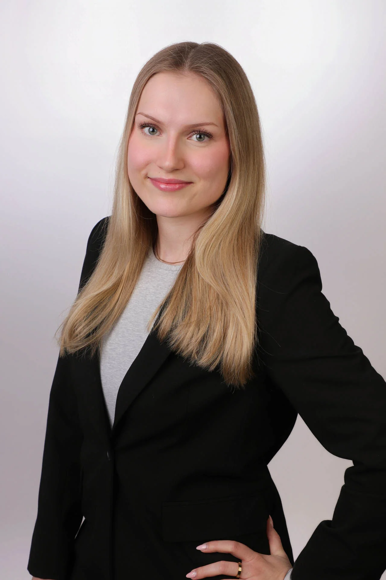 Eine Frau mit glattem blondem Haar trägt einen schwarzen Blazer und ein hellgraues Oberteil, steht vor einem schlichten hellen Hintergrund und lächelt in die Kamera.
