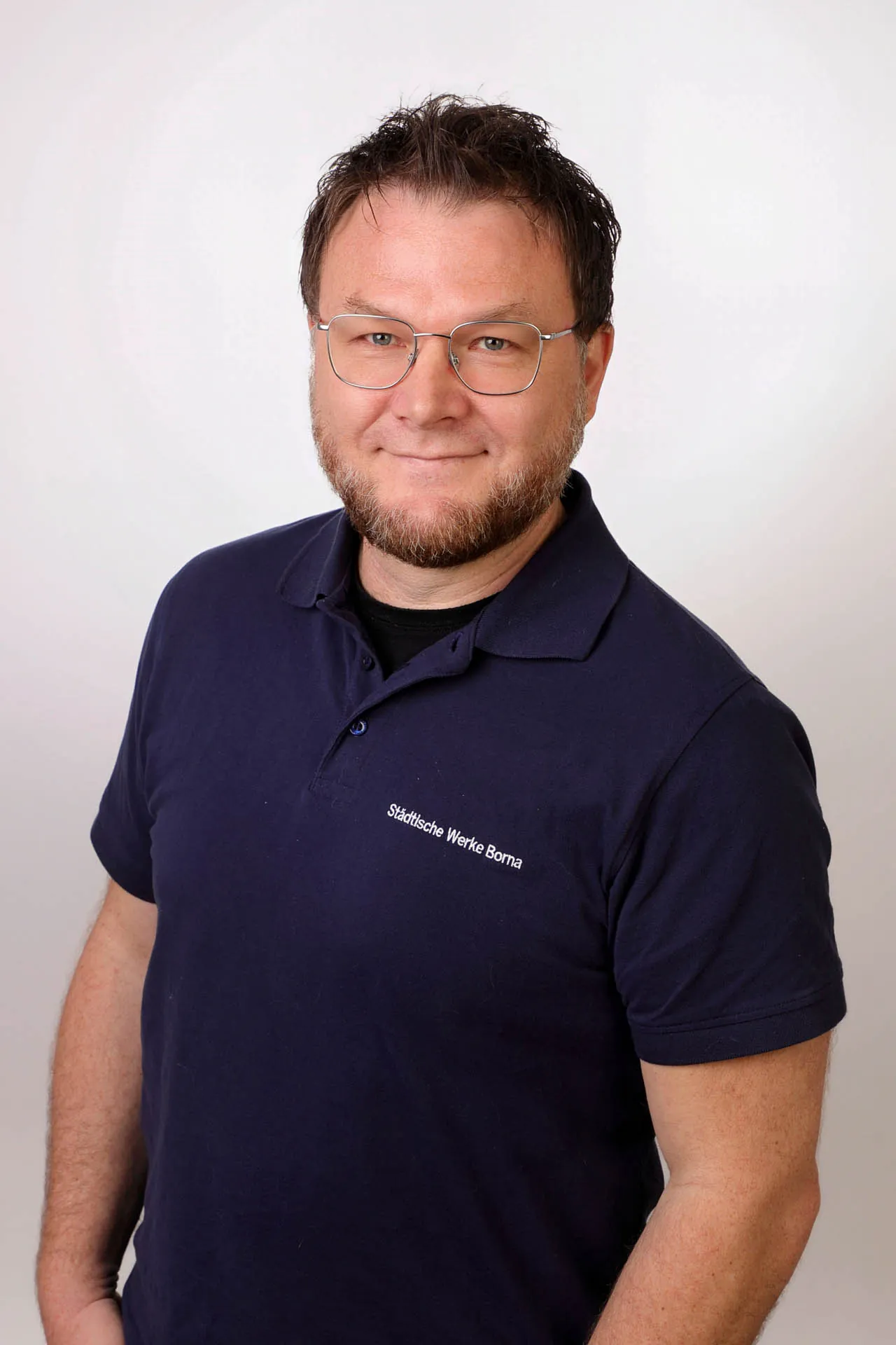 Ein Mann mit Brille und kurzem Bart trägt ein marineblaues Polohemd und steht vor einem schlichten, hellen Hintergrund, mit neutralem Blick in die Kamera.
