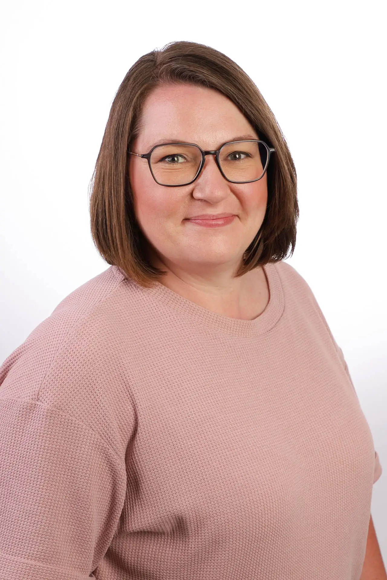 Eine Frau mit kurzen braunen Haaren und Brille, die ein hellrosa strukturiertes Oberteil trägt, posiert vor einem schlichten weißen Hintergrund.